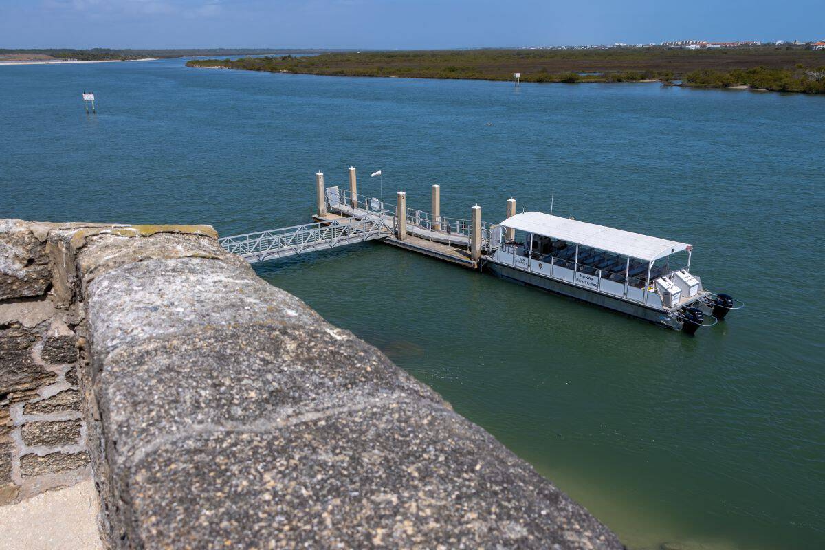 A white passenger boat is docked at a floating pier on a wide river, viewed from behind a stone wall. Trees and buildings are visible in the distance.