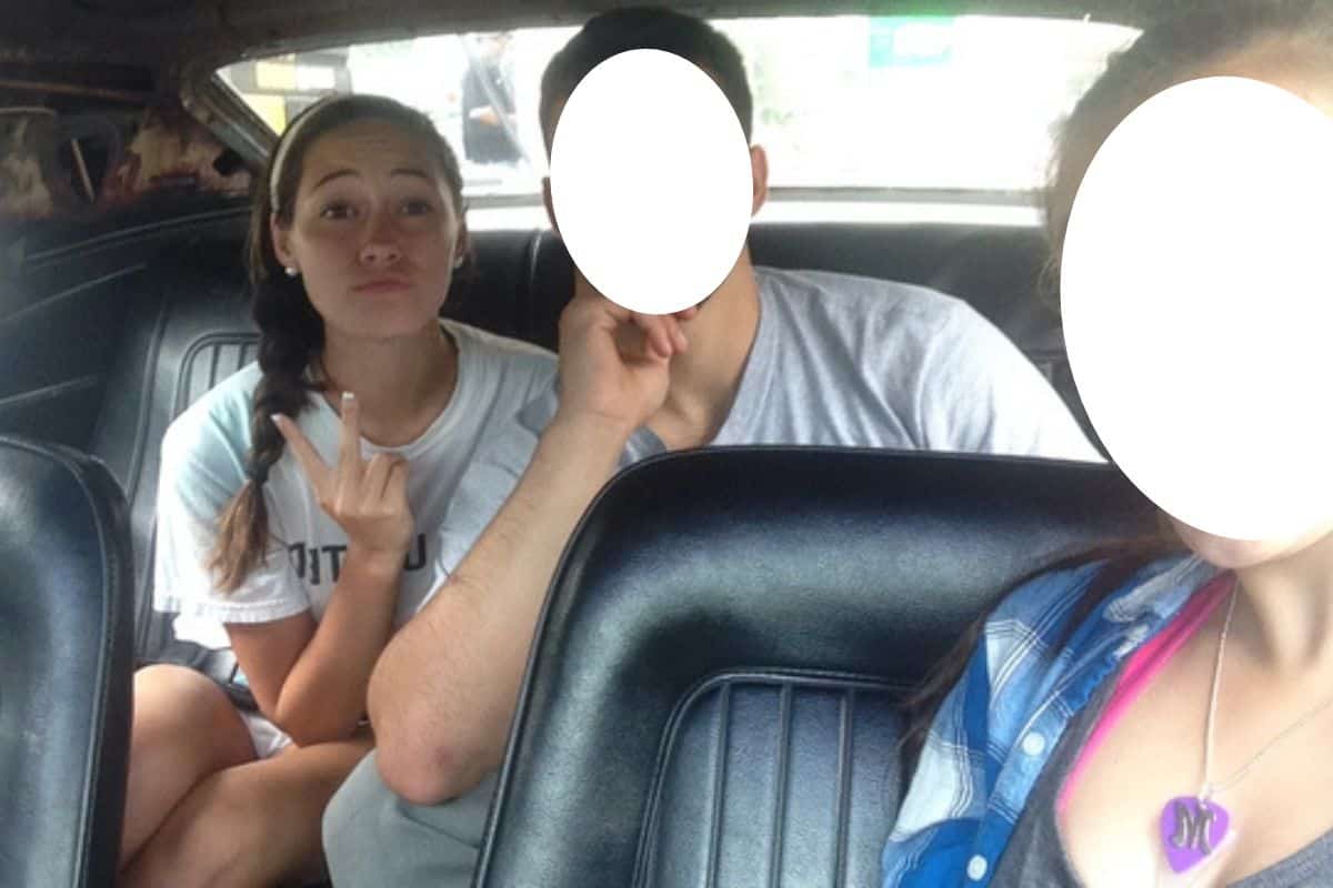 Three people sitting in the backseat of a car; the faces of two individuals are blurred, while the third person makes a peace sign.