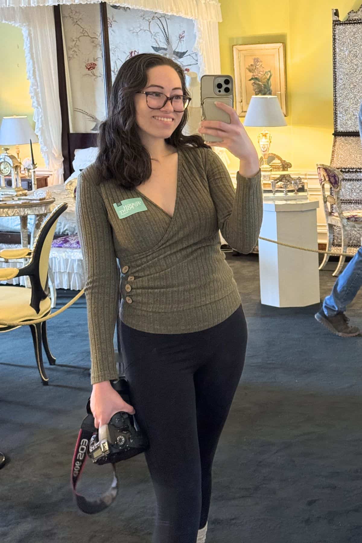 A woman with glasses takes a mirror selfie in a room with antique furniture, holding a camera in one hand and a smartphone in the other.