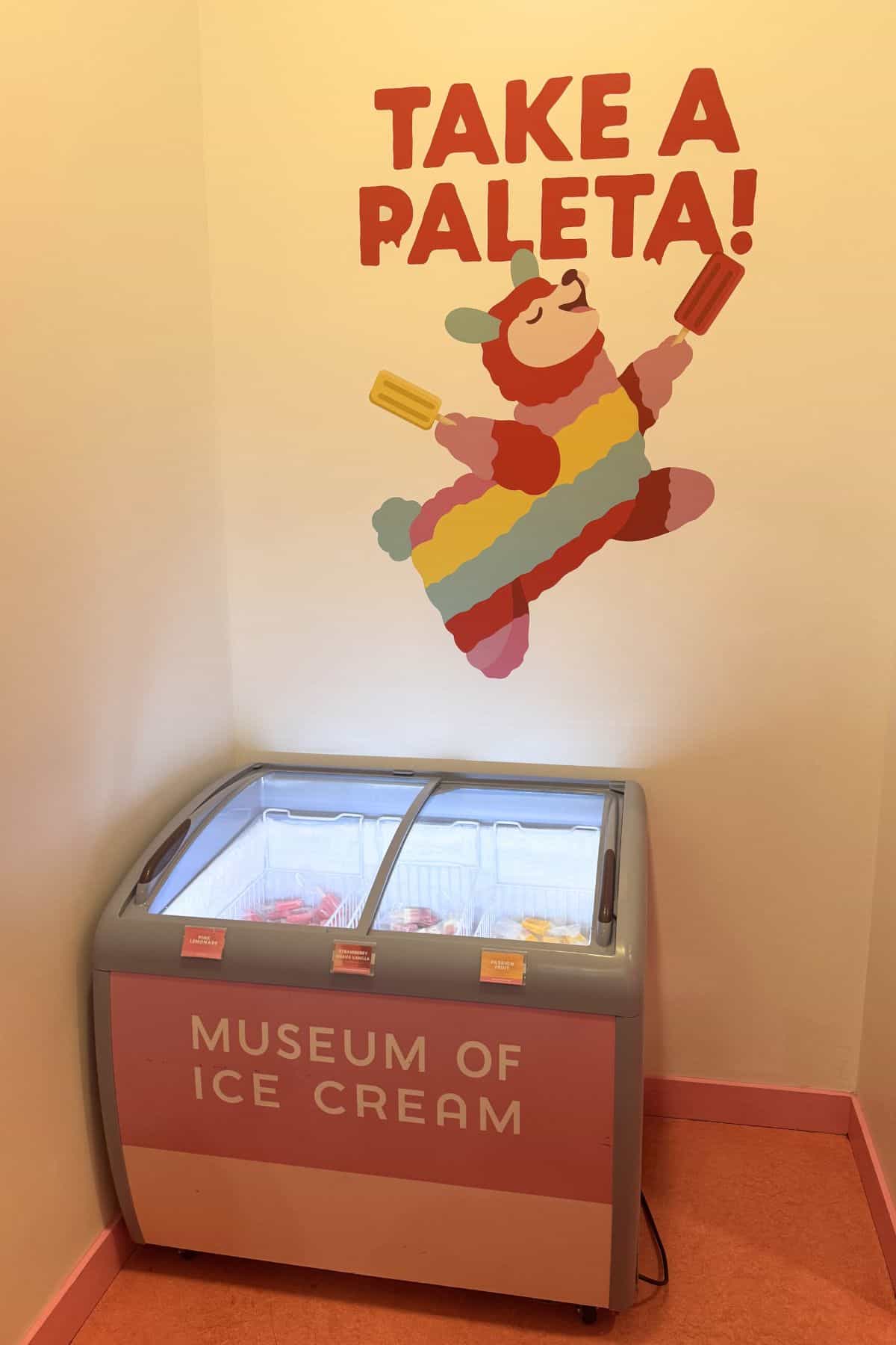 Chest freezer labeled "Museum of Ice Cream" below a mural of a bear in a rainbow outfit holding popsicles with the text "Take a Paleta!" on the wall.
