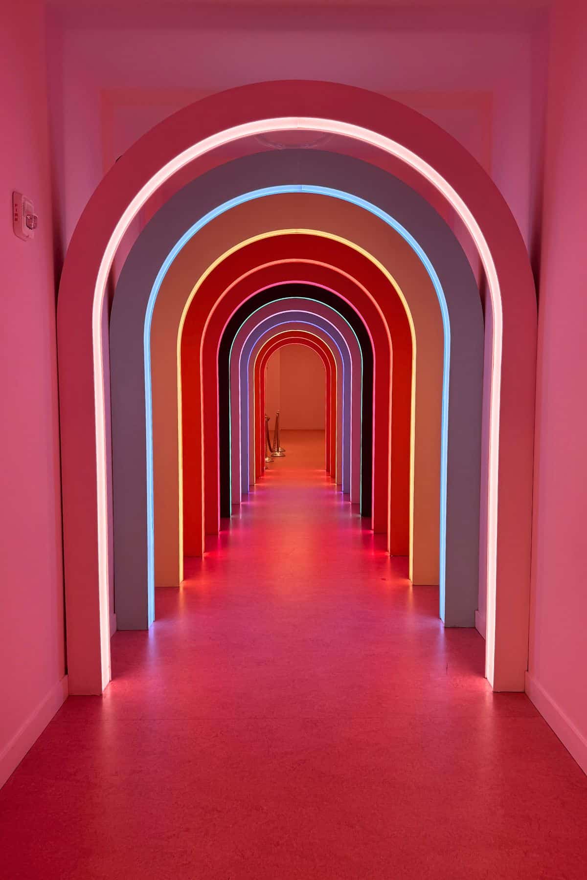 A hallway with a series of illuminated, colorful archways in red, pink, and blue, creating a tunnel effect with glowing lights on the floor and walls.