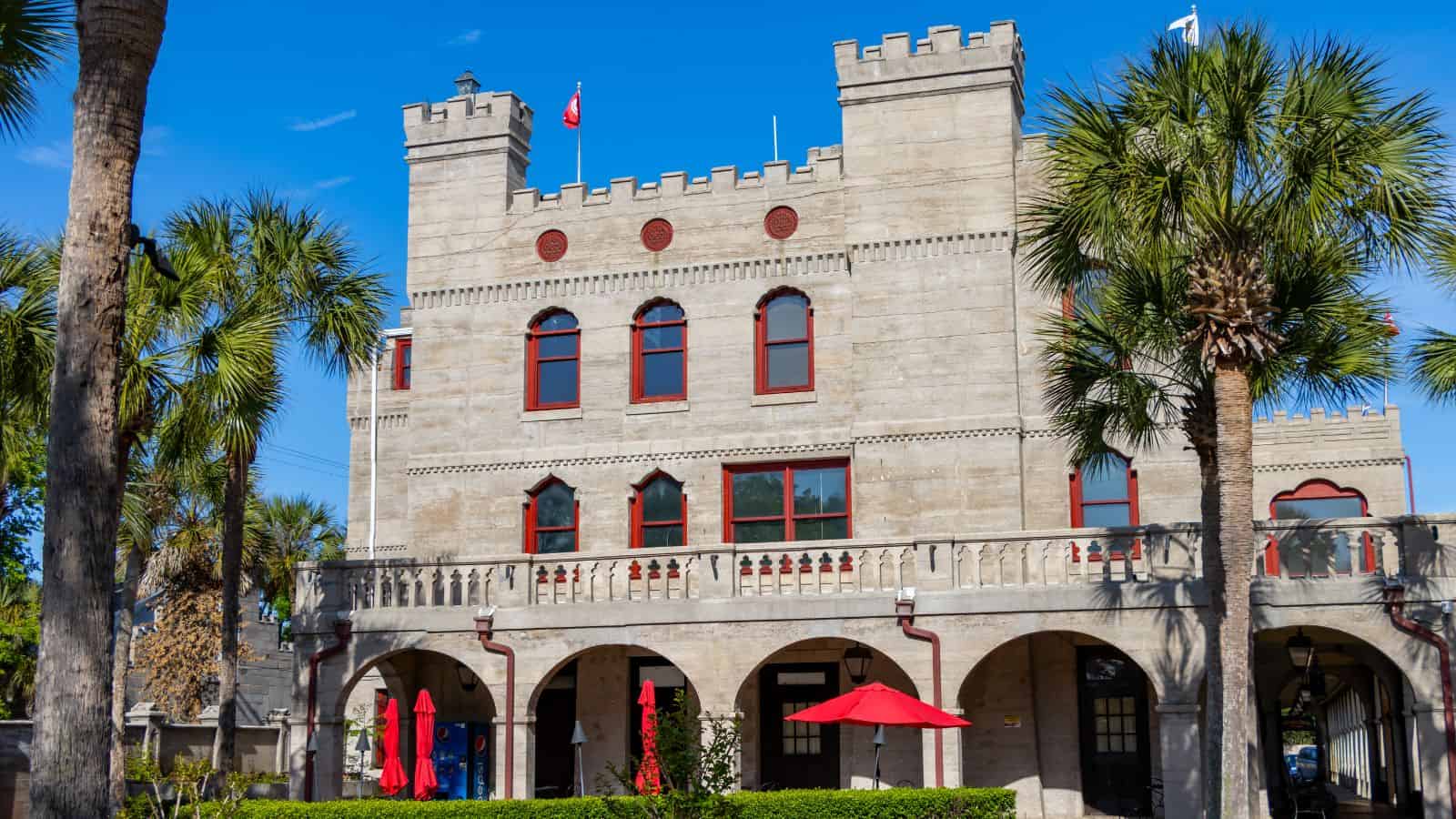 A stone building with castle-like features, including battlements and arched windows, stands surrounded by palm trees. Red umbrellas and patio furniture are visible in front.
