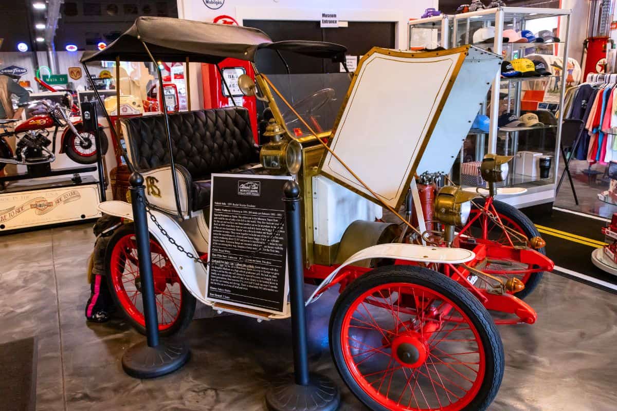 A vintage white and red automobile with an open hood is displayed indoors, surrounded by informational signage and souvenirs in the background.