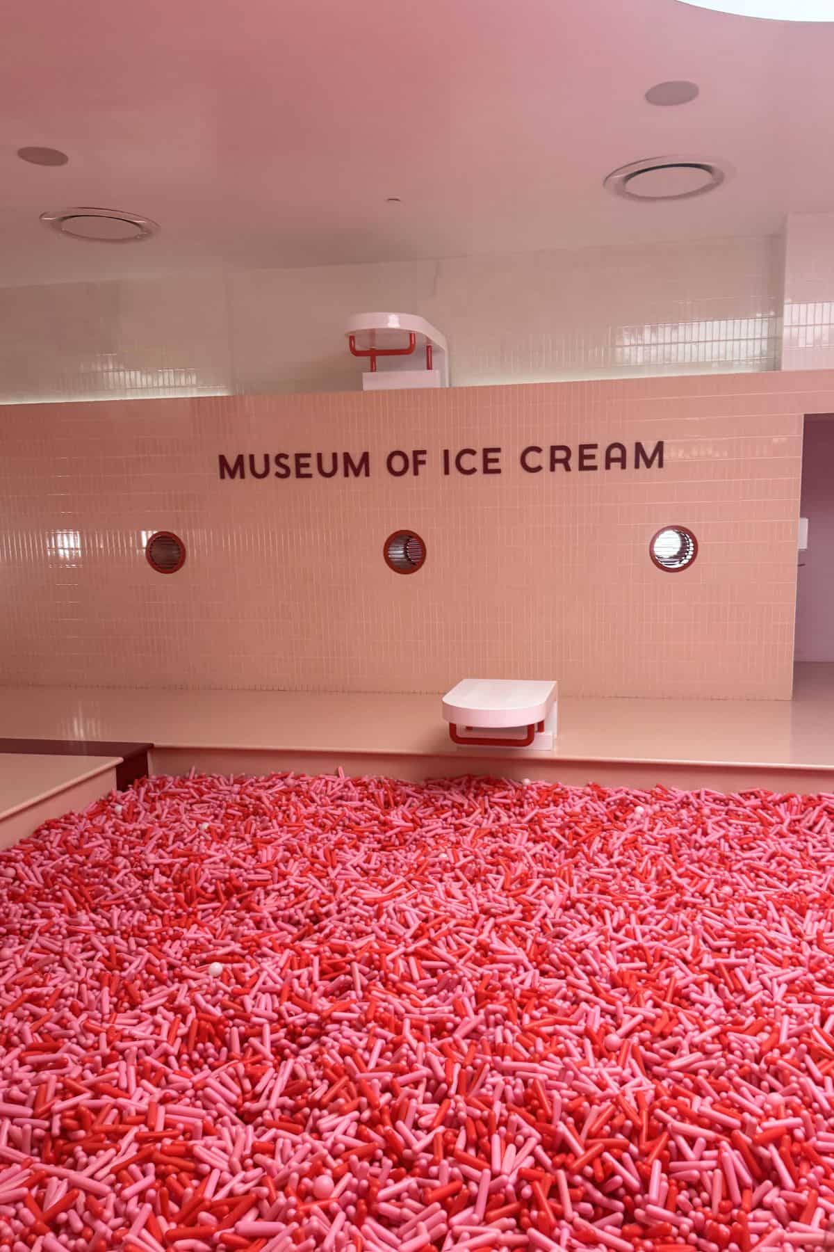 A pink-themed room at the Museum of Ice Cream with a pool filled with red and pink sprinkles and a white wall displaying the museum's name.
