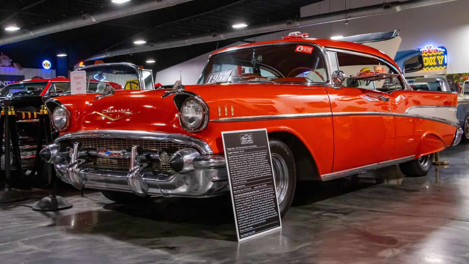 A red 1957 Chevrolet Bel Air is displayed indoors at a car show, with an informational sign in front of the vehicle.