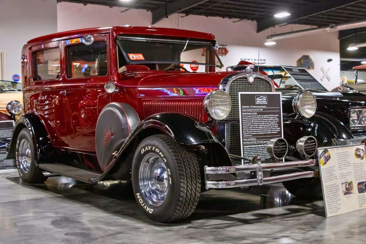A vintage maroon and black four-door car is displayed indoors with informational signs in front of it, surrounded by other classic cars.