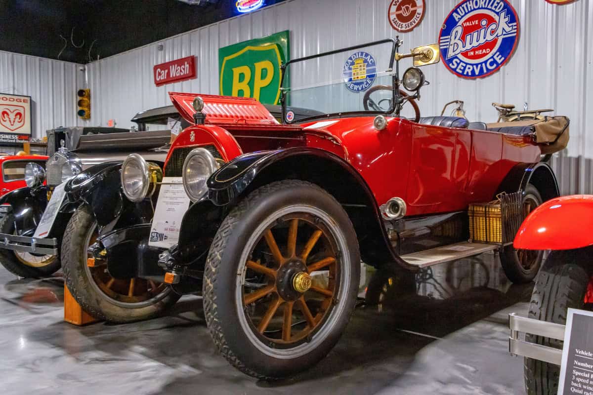 A vintage red convertible car with wooden-spoke wheels is displayed indoors, surrounded by automotive signs and other classic cars.