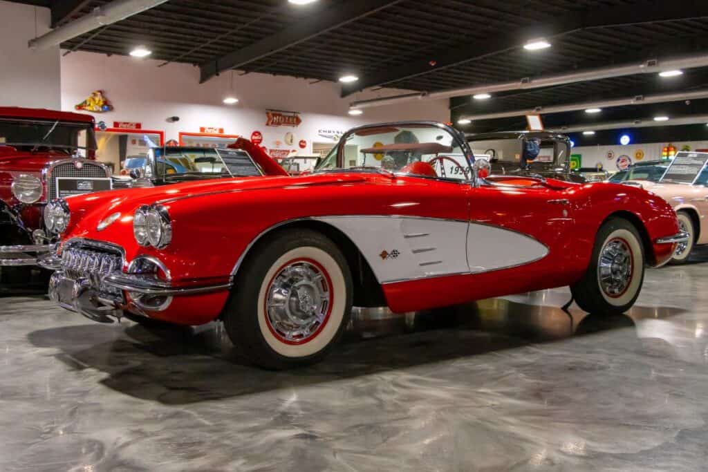 A classic red and white convertible car is displayed indoors among other vintage vehicles under bright overhead lighting.