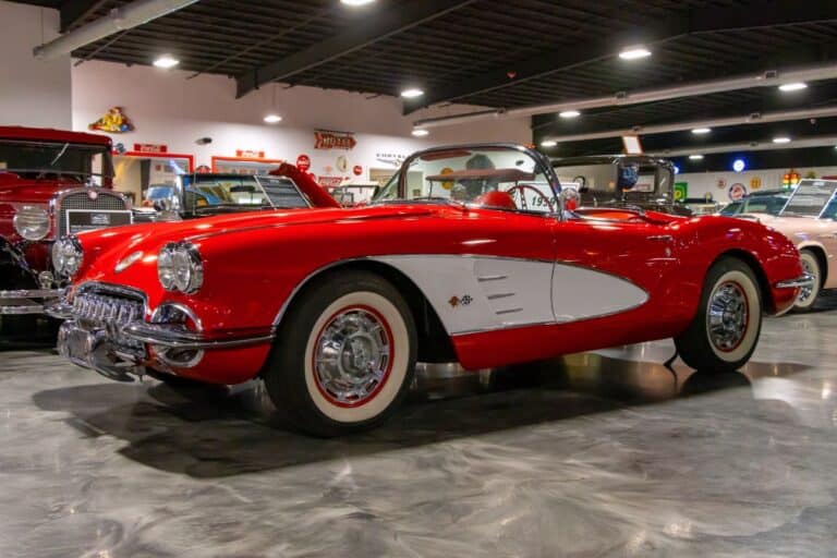 A classic red and white convertible car is displayed indoors among other vintage vehicles under bright overhead lighting.