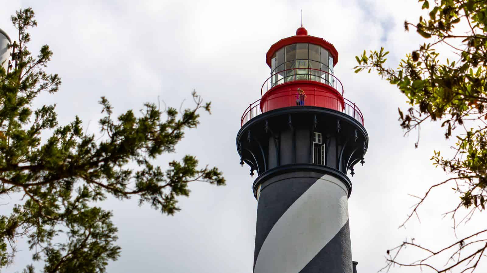 A black and white striped lighthouse with a red top is surrounded by tree branches against a cloudy sky.