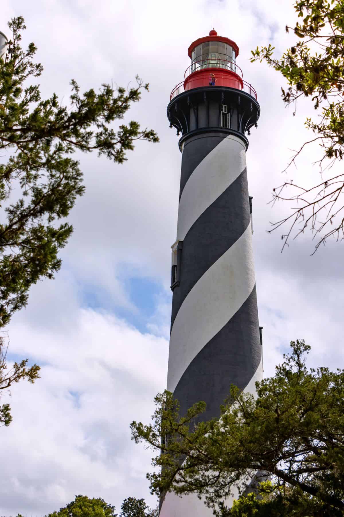 A tall lighthouse with black and white spiral stripes and a red top stands among trees under a cloudy sky at Fort Matanzas National Monument.