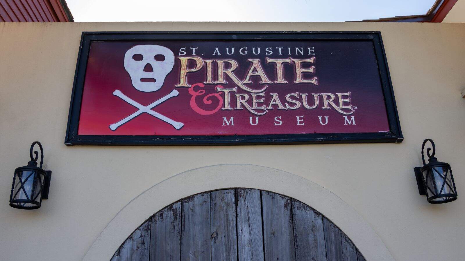 Sign for the St. Augustine Pirate & Treasure Museum with a skull and crossbones, flanked by two lanterns above a wooden arched door.