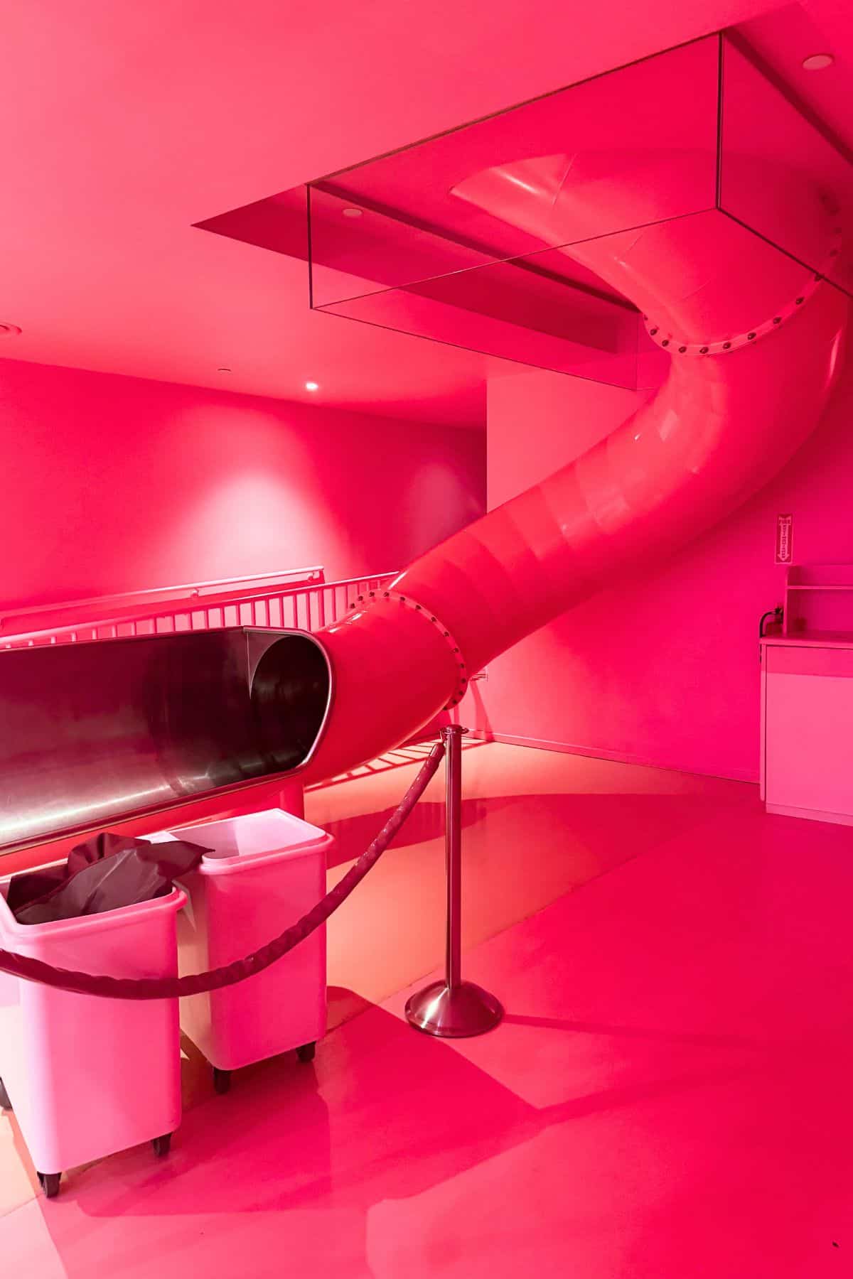 A large pink indoor slide curves down from the ceiling, ending near two white bins and a rope barrier in a brightly lit pink room.
