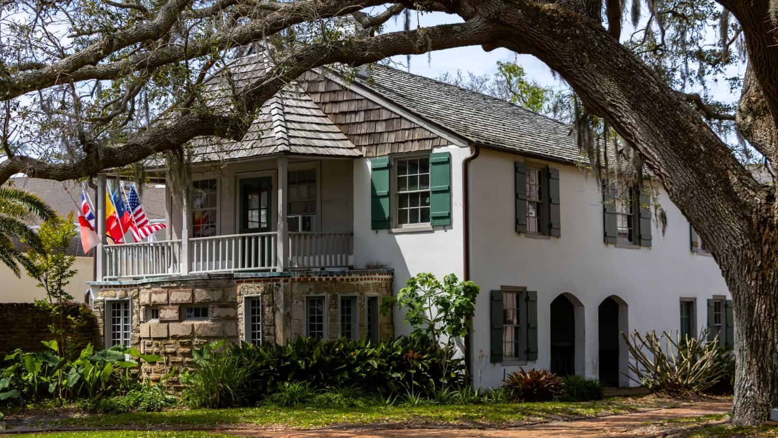 A historic two-story white house with green shutters, multiple flags, a stone porch, arched doorways, and large oak trees in front.