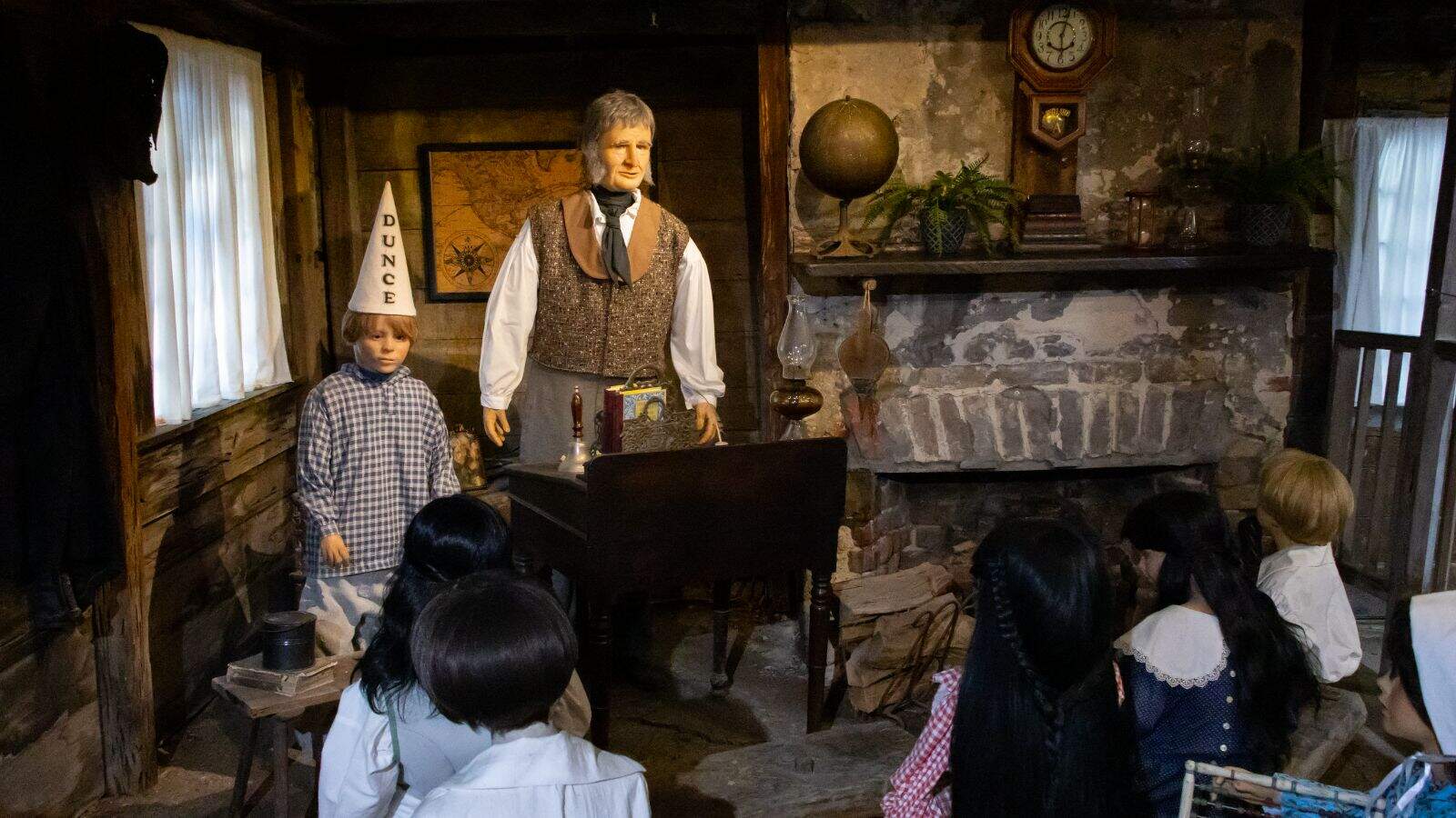 A historical classroom scene with mannequins, featuring a teacher, students, and a child wearing a dunce cap standing at the front near the teacher’s desk.