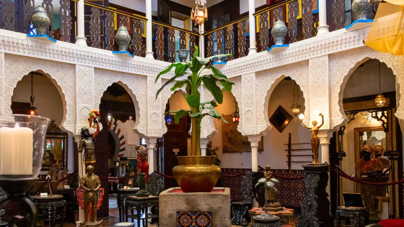Ornate interior courtyard with carved arches, intricate woodwork, decorative lanterns, a potted plant in the center, and various sculptures and artifacts displayed around the space.