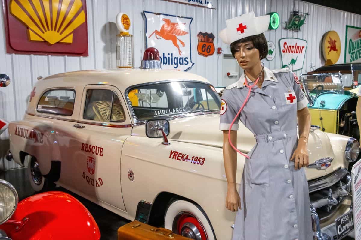 A mannequin dressed as a nurse stands next to a vintage rescue squad ambulance in a museum with various retro signs displayed on the wall.