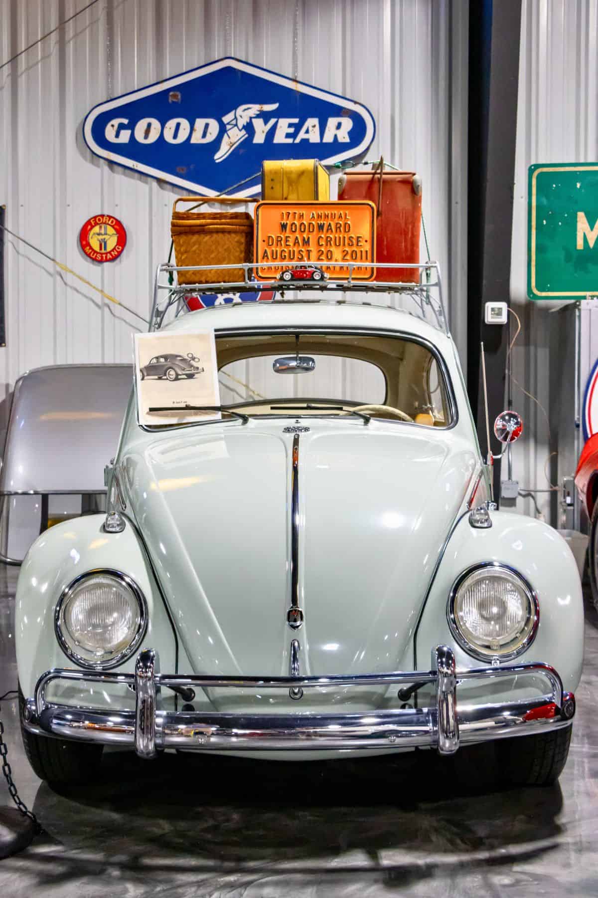Front view of a vintage light blue Volkswagen Beetle with a roof rack carrying baskets and a sign, displayed indoors near a Goodyear sign.