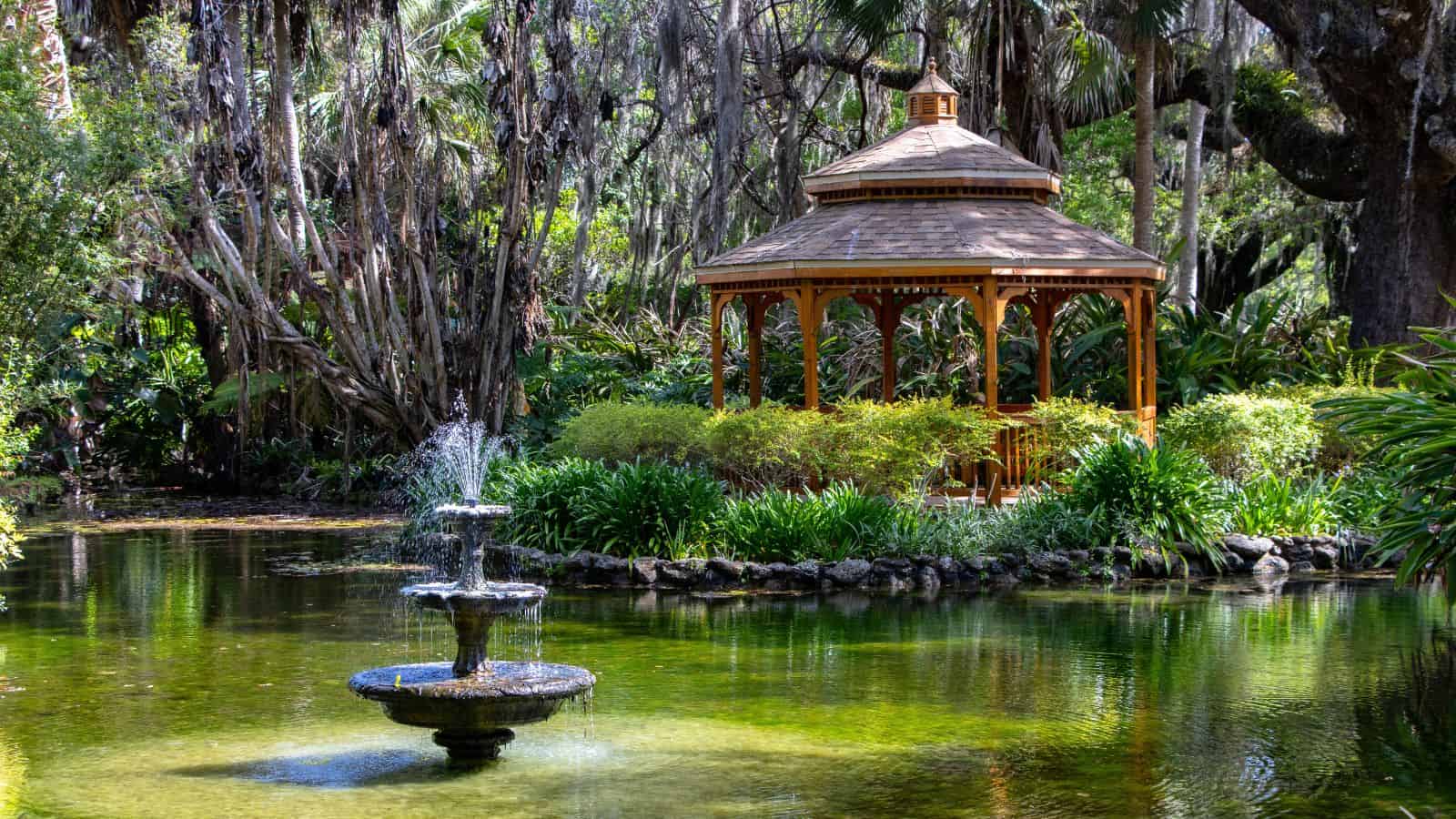 A wooden gazebo stands near a pond with a tiered fountain in a lush garden surrounded by trees and greenery.