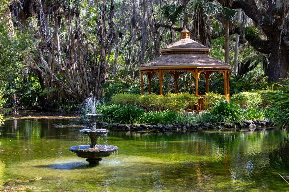 A wooden gazebo stands surrounded by lush greenery and trees at Fort Matanzas National Monument, with a tiered water fountain in a pond in the foreground.