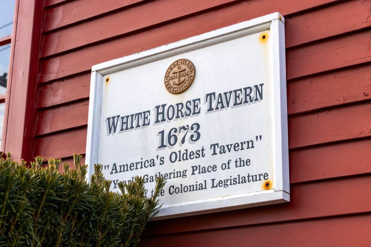 A sign on a red wooden exterior reads "White Horse Tavern 1673, America's Oldest Tavern, Meeting Place of the Colonial Legislature." A bush is partially visible in the foreground.