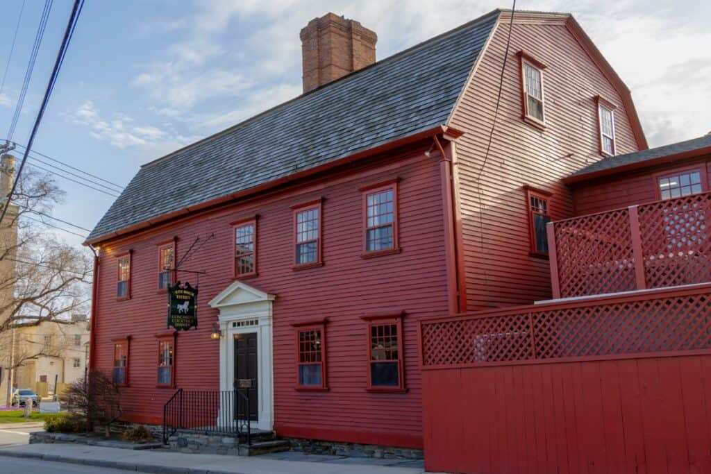 A large red wooden colonial-style house with a central chimney, white trim, and a fenced patio, located on a quiet street under a partly cloudy sky.