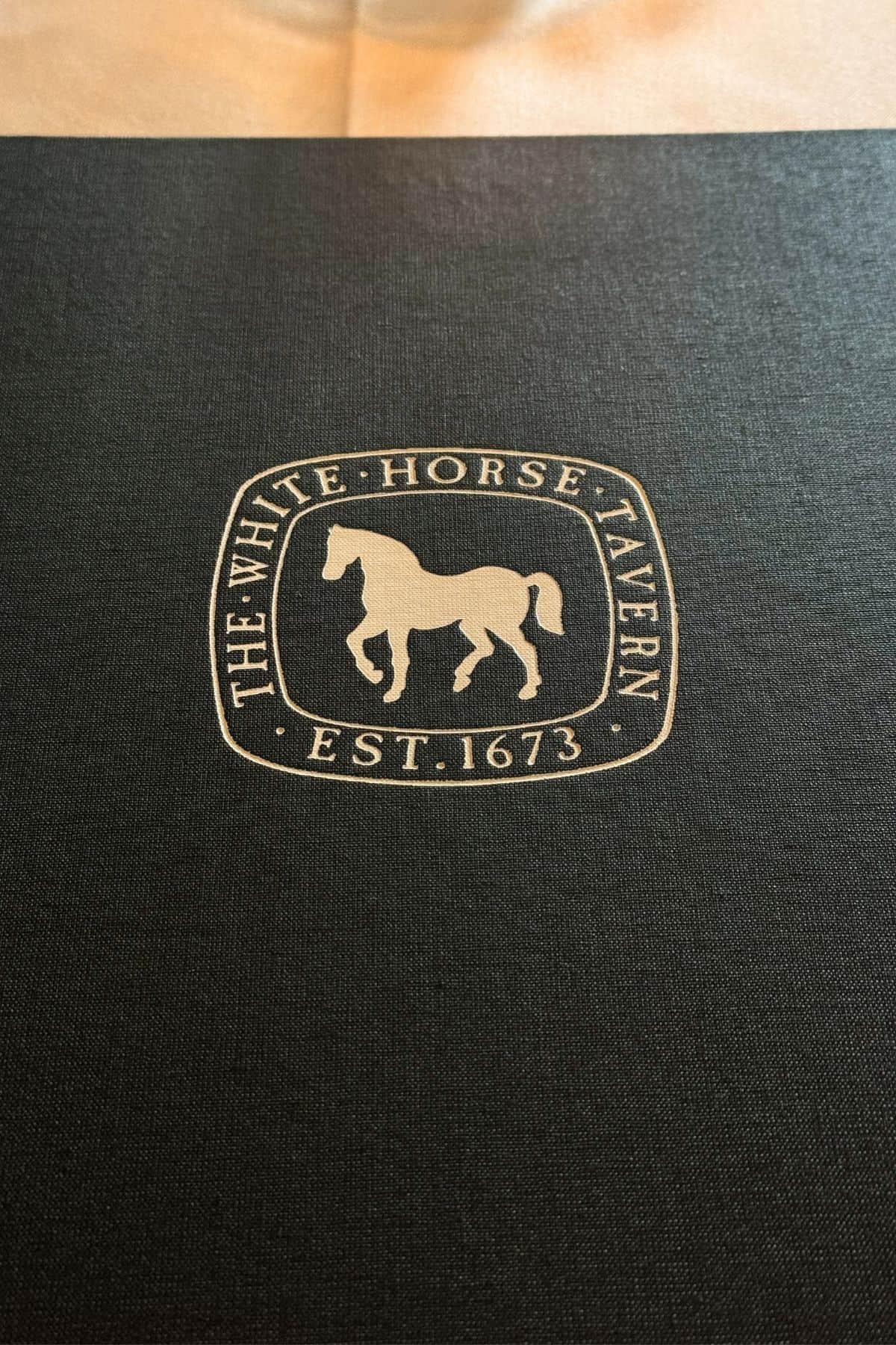 A close-up of the White Horse Tavern emblem featuring a horse and the text “THE WHITE HORSE TAVERN EST. 1673” on a dark background.