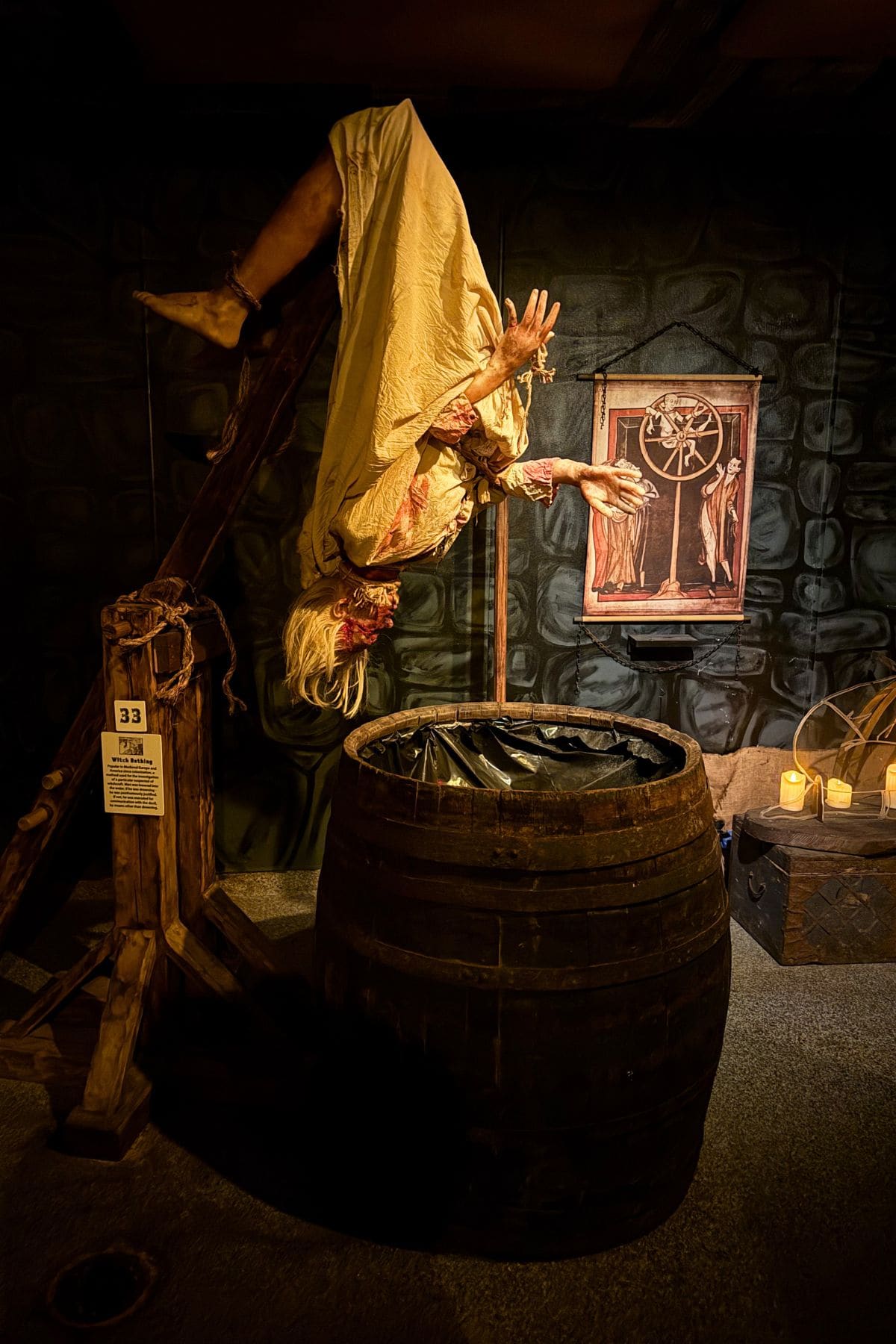 A mannequin dressed as a distressed woman hangs upside down over a wooden barrel in a dim, medieval-themed room with candles and torture devices.