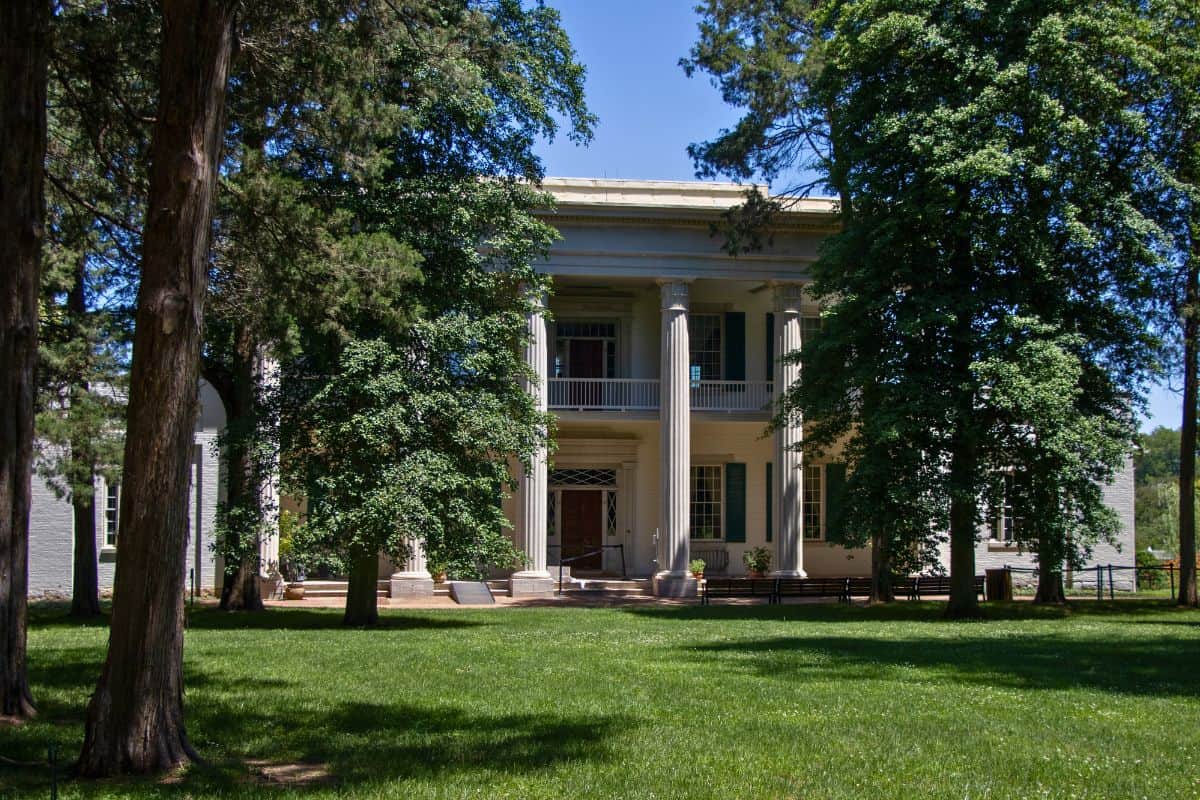 Large white two-story mansion with columns, partially obscured by tall trees, sits on a well-kept green lawn under a clear blue sky.