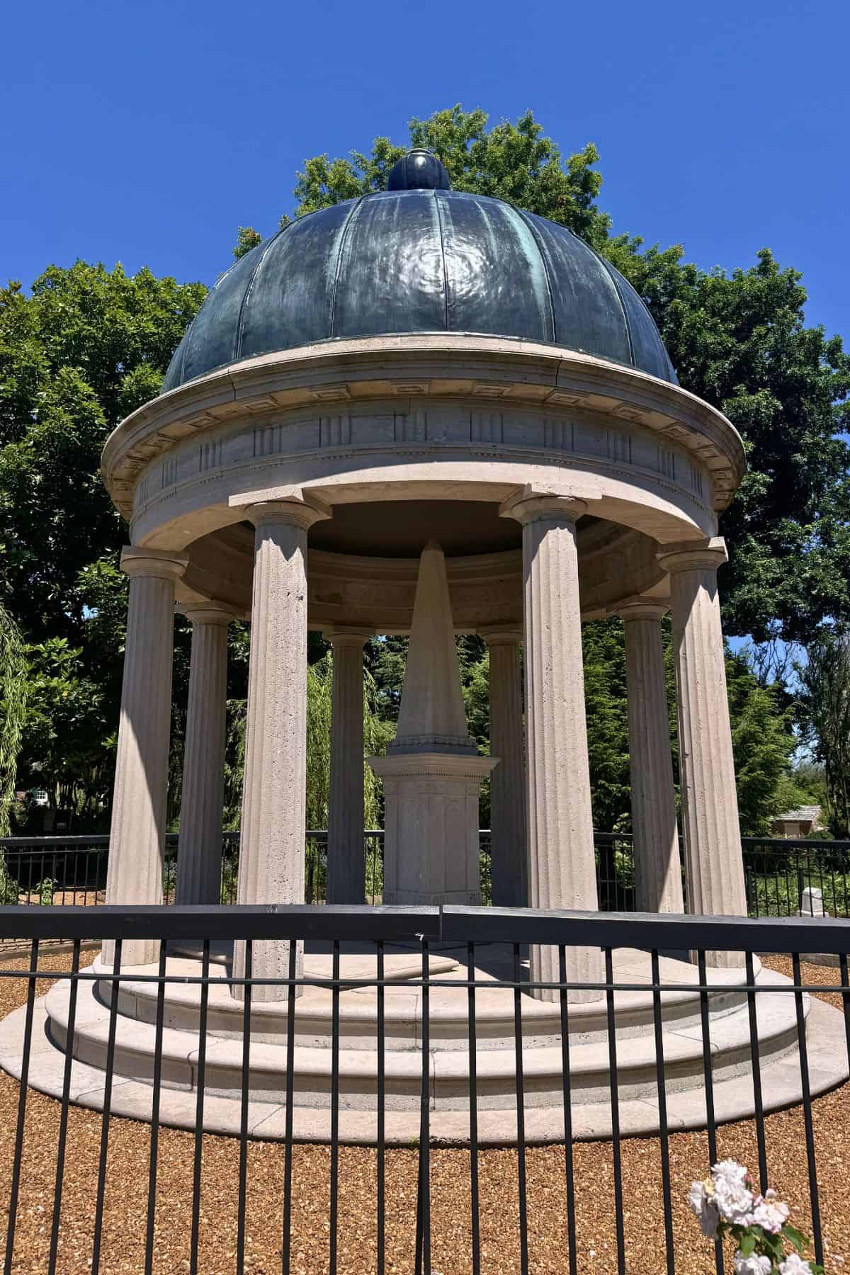 A domed stone pavilion with columns surrounds an obelisk monument, set in a landscaped area with trees and a metal fence in the foreground.
