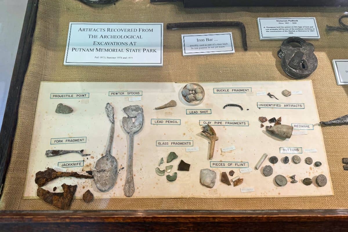 A display case containing labeled artifacts from archaeological excavations, including flint, glass, metal objects, buttons, stones, and pottery fragments.