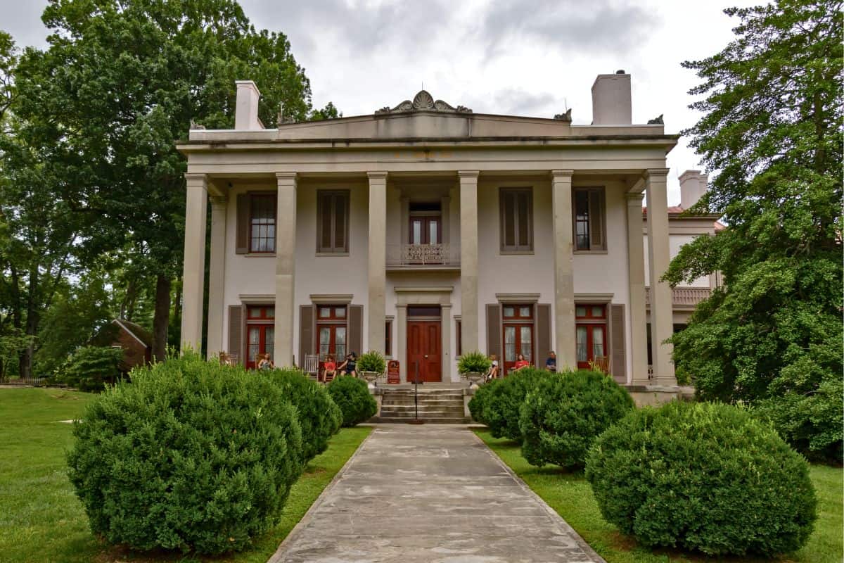A large, two-story historic mansion with tall columns, symmetrical windows, and a manicured lawn with trimmed bushes leading to the entrance.