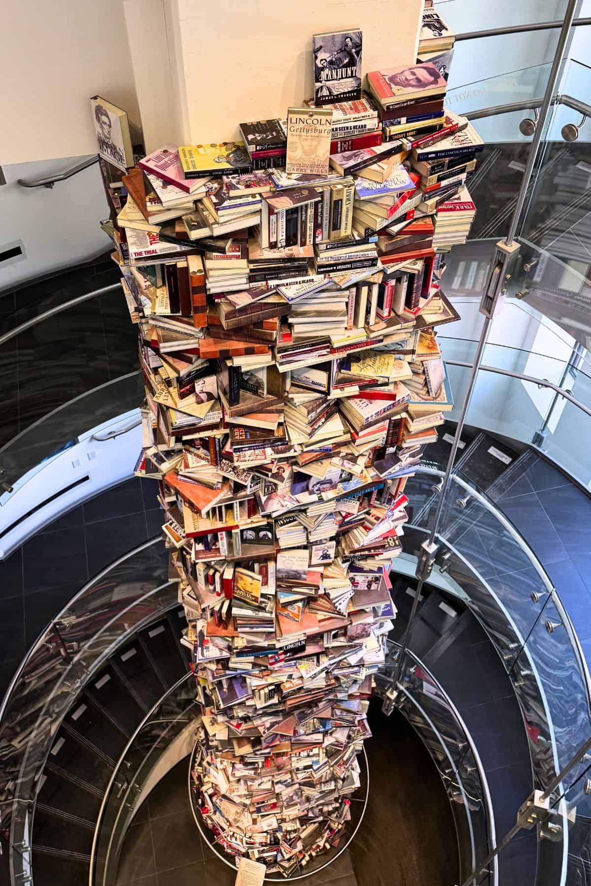 A tall, cylindrical stack of books stands in the center of a spiral staircase in a modern building with glass railings.