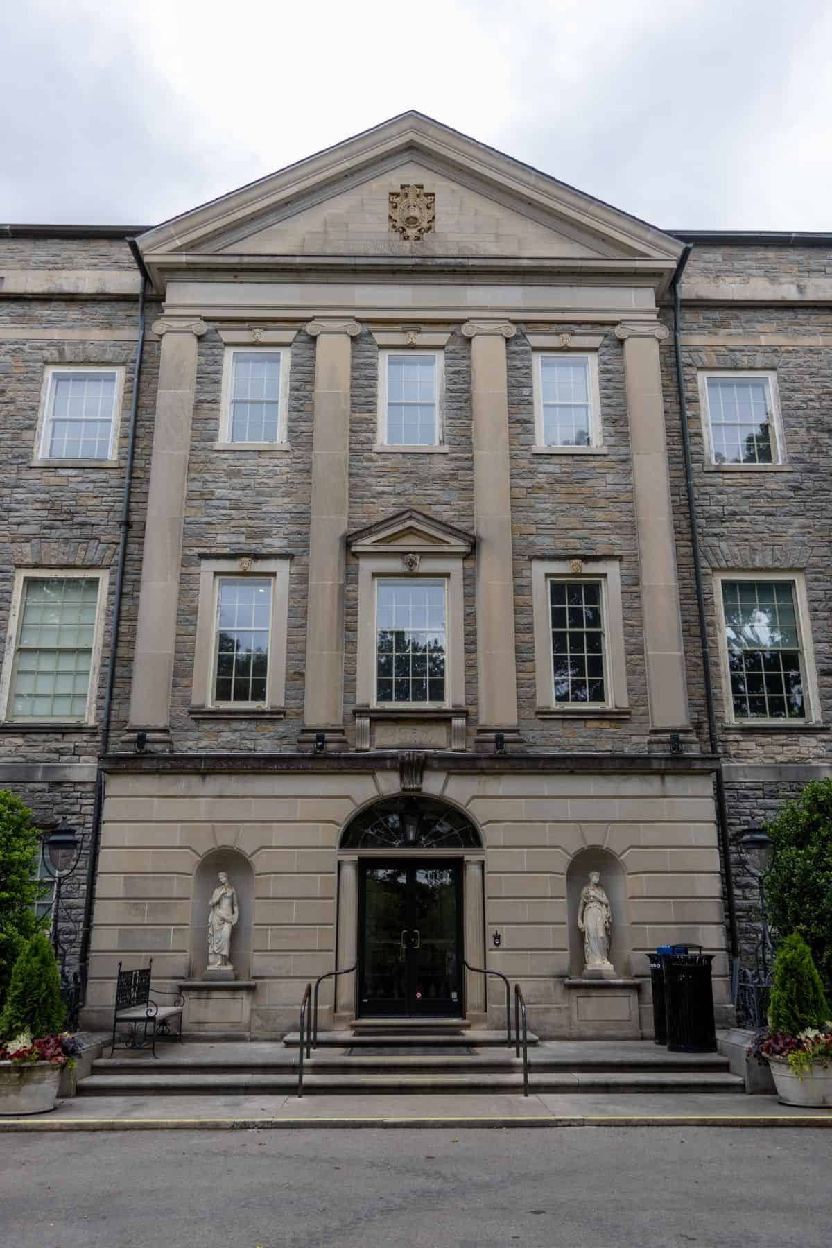 Three-story stone building with columns, arched entrance, two statues by the doorway, and symmetric windows; cloudy sky in the background.