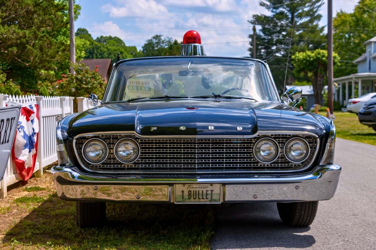 Front view of a vintage black Ford police car with a red light on top, parked on a residential street. The license plate reads "BULLET.