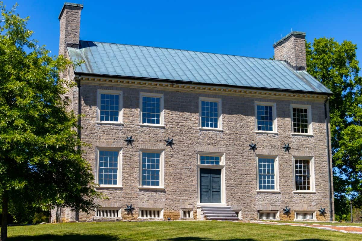 A two-story stone house with a metal roof, symmetrical windows, and a central front door, surrounded by trees and grass under a clear blue sky.