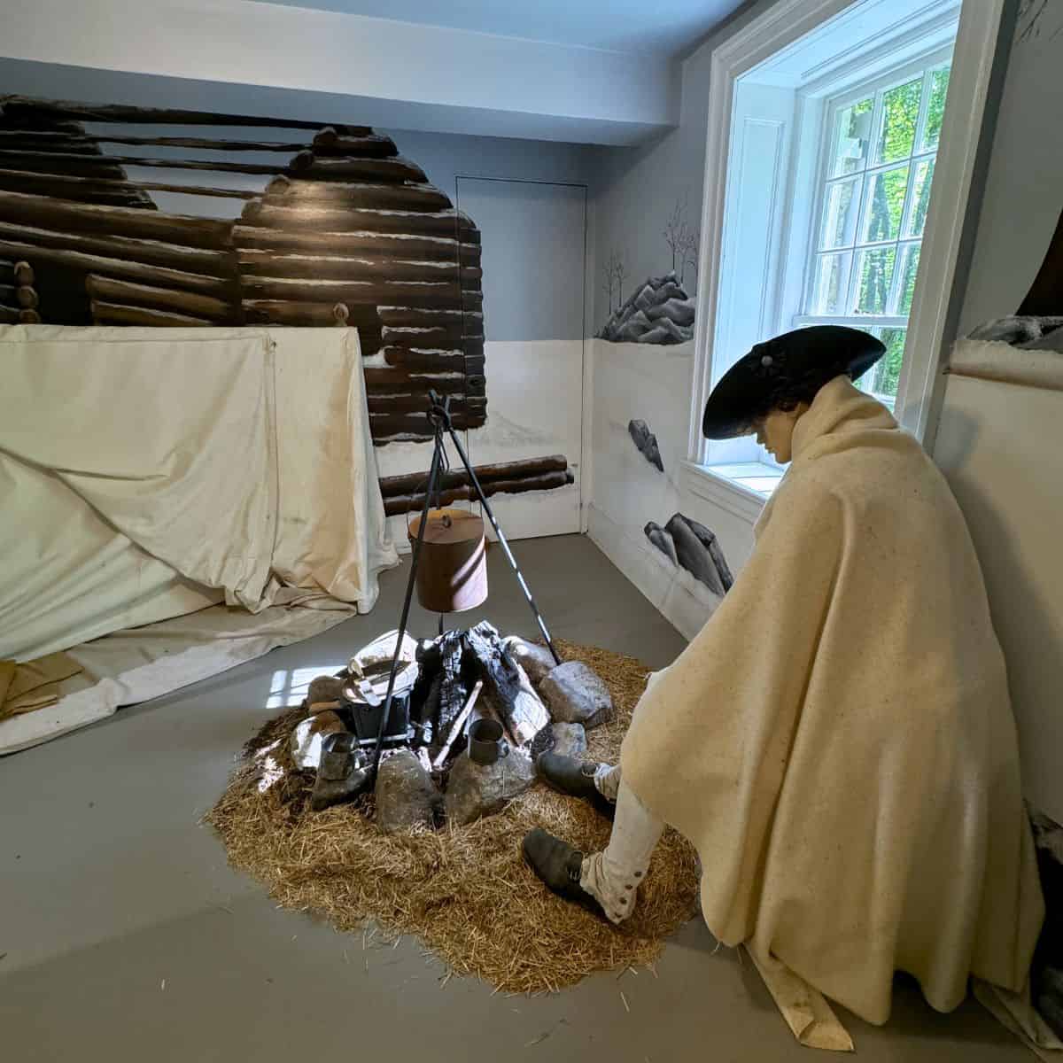 A mannequin dressed in colonial attire sits by a fake campfire with a pot, inside a room with a log cabin backdrop and a large window.