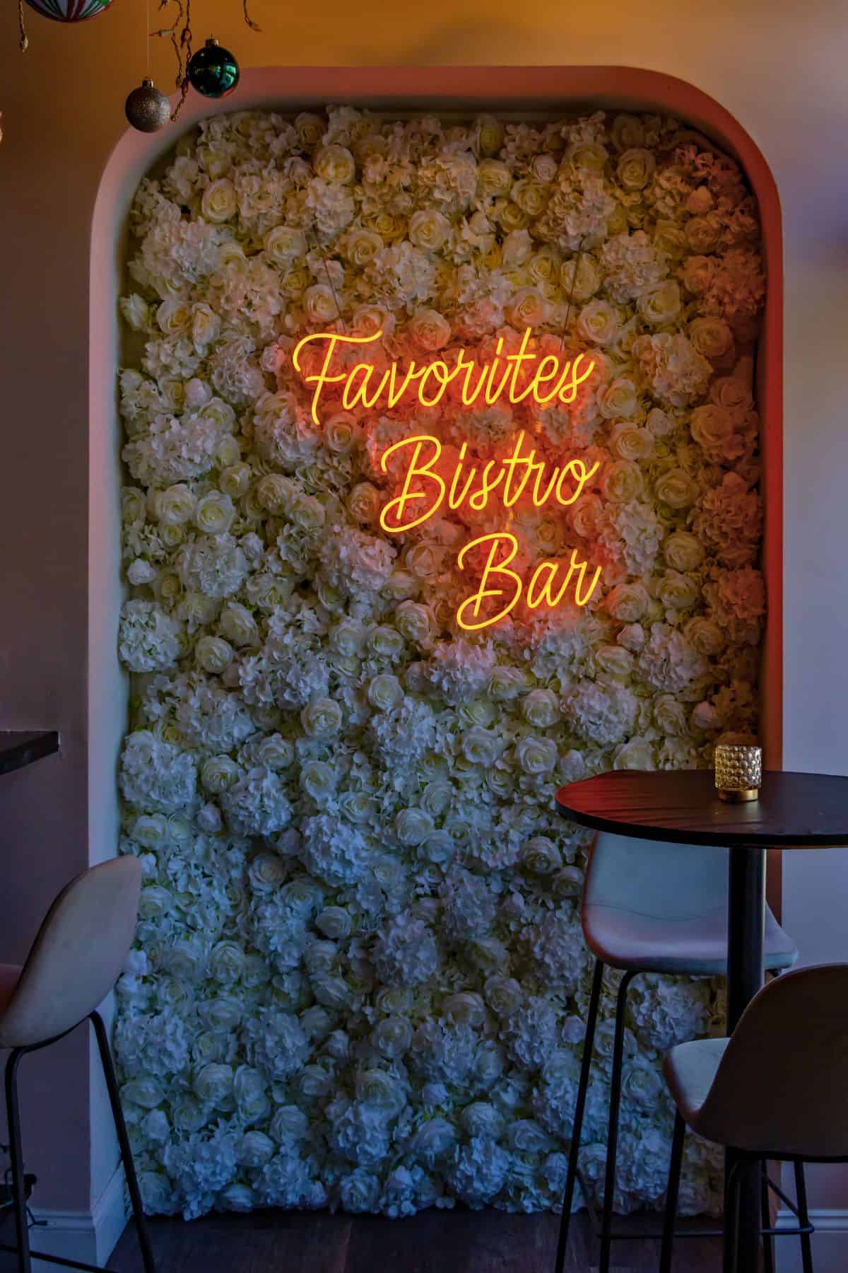 Neon sign reading "Favorites Bistro Bar" on a wall covered with white artificial flowers, next to a small round table and two chairs.
