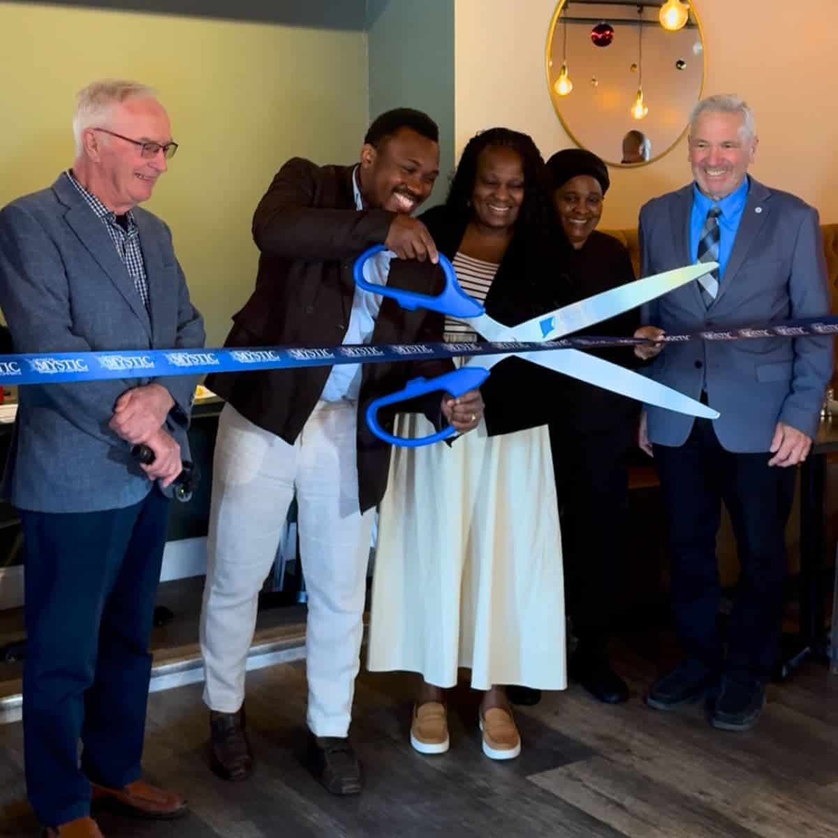Five people stand together at a ribbon-cutting ceremony, with two holding oversized scissors and cutting a blue ribbon indoors.