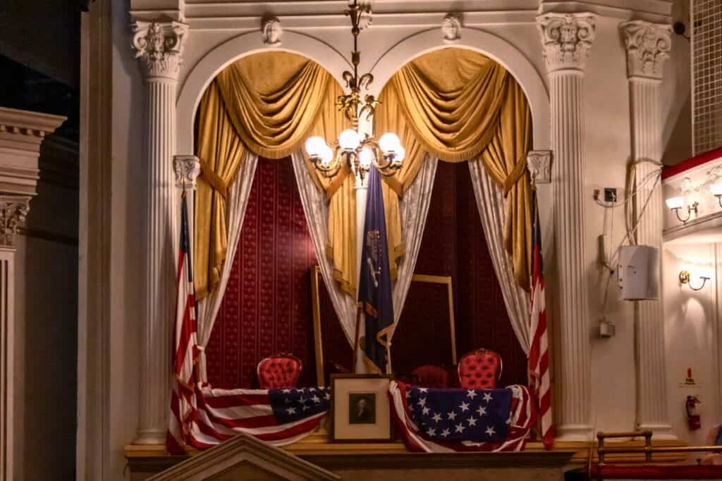 Theatre box draped with gold curtains and American flags, containing two ornate red chairs and a framed portrait, with columns and a chandelier above.