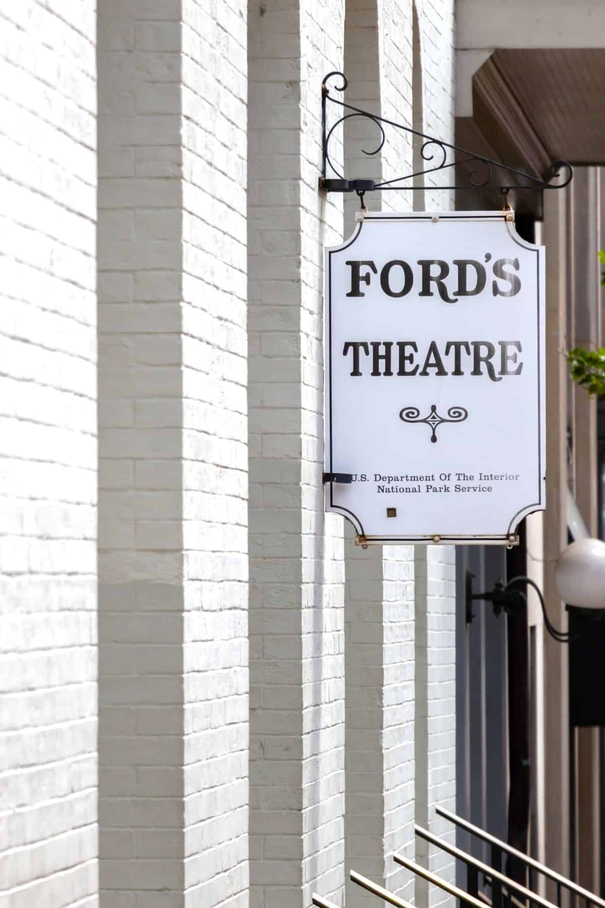 A white sign with black text reading “Ford’s Theatre” hangs on a white brick building. Smaller text below mentions the U.S. Department of the Interior and the National Park Service.