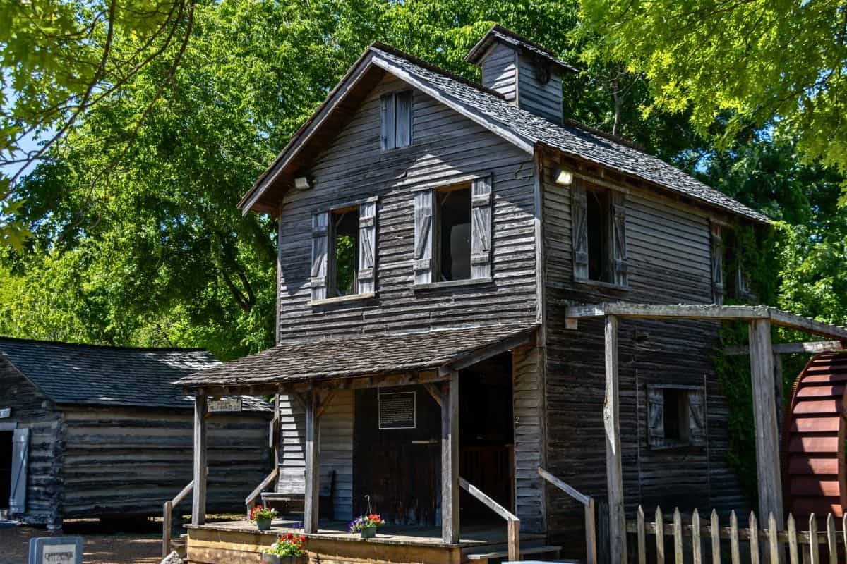 A two-story wooden building with weathered siding, open windows, and a small covered porch, surrounded by trees and other rustic structures.