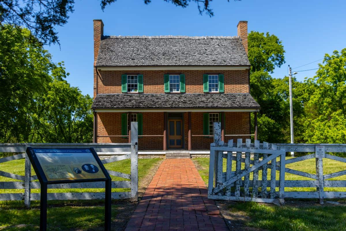 A historic two-story brick house with green shutters sits behind a white wooden fence and open gate, with an informational sign in the foreground and trees in the background.