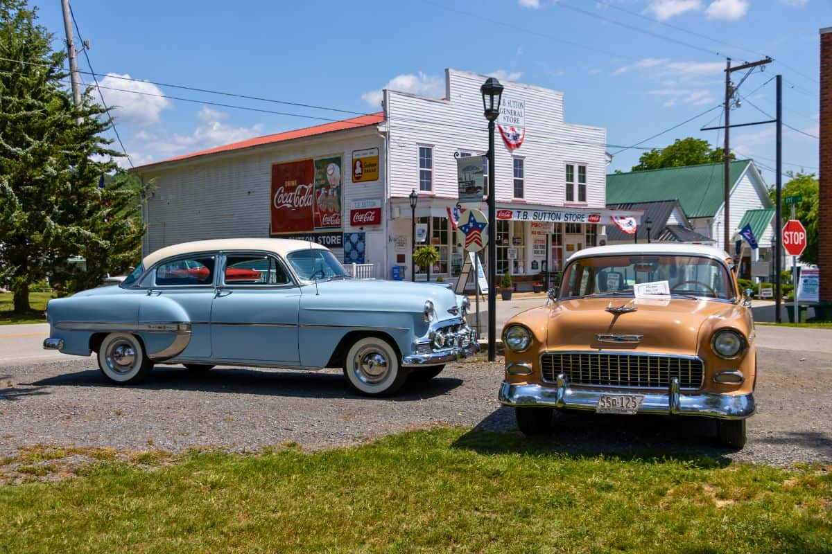 Two vintage cars are parked on a street in front of an old-fashioned general store with Coca-Cola signage on a sunny day.