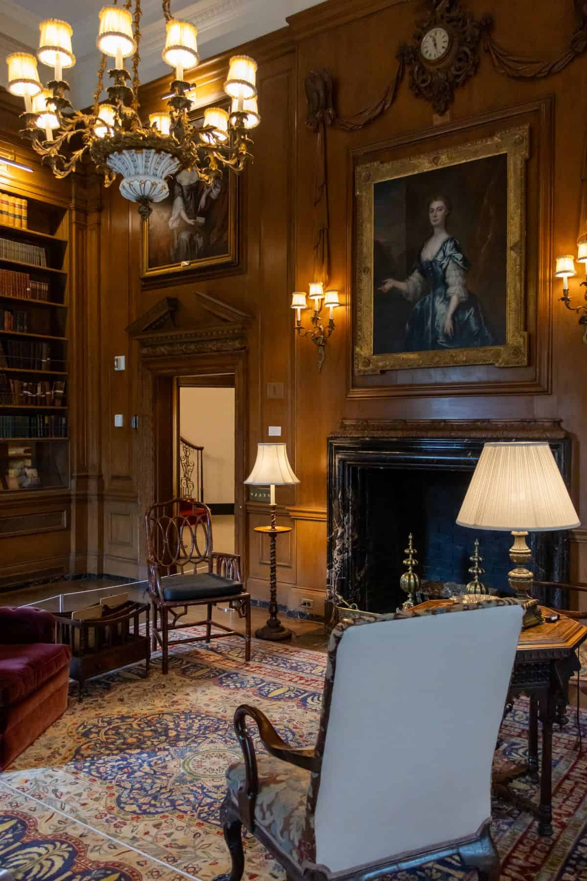 Elegant library with wood-paneled walls, ornate chandelier, large fireplace, antique furniture, bookshelves, and classical portraits above the mantelpiece.