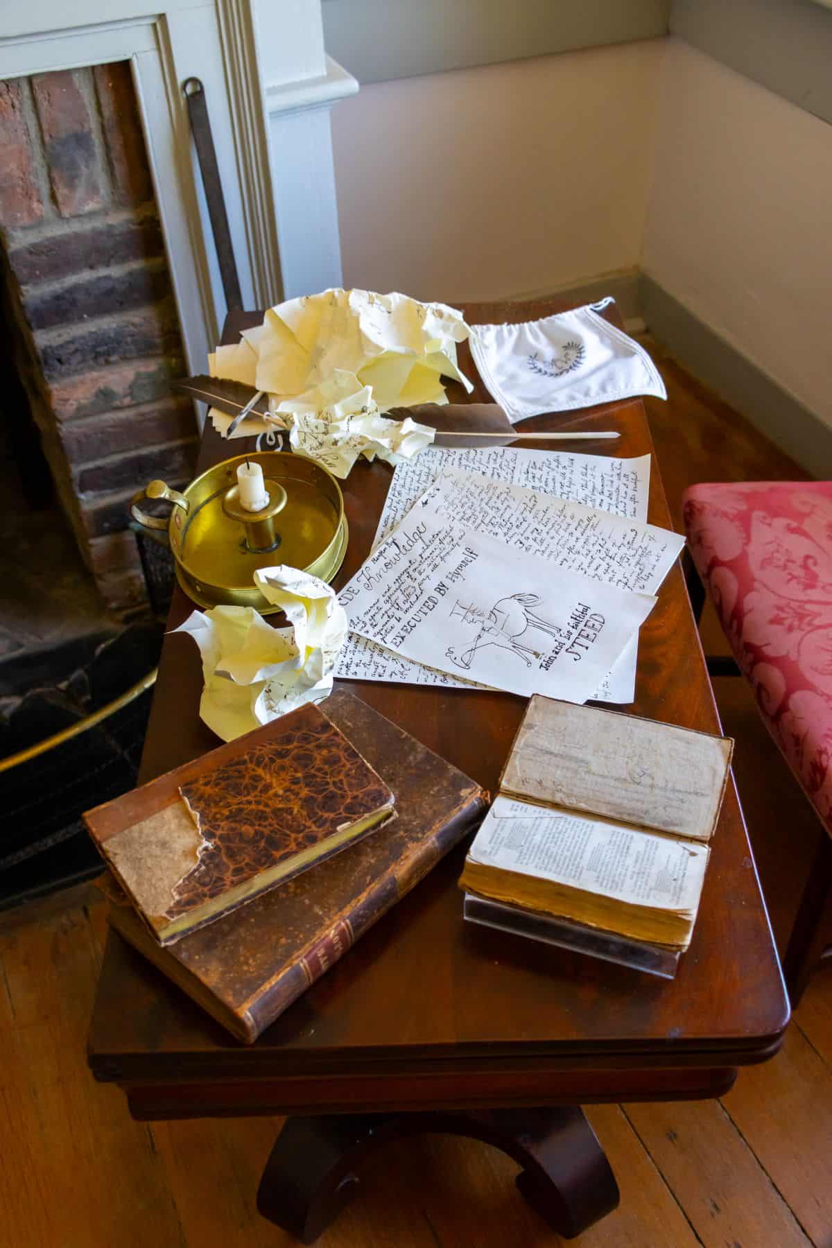 A wooden table with old books, handwritten notes, crumpled papers, a candlestick, and a folded white cloth, set beside a fireplace and a pink upholstered chair.