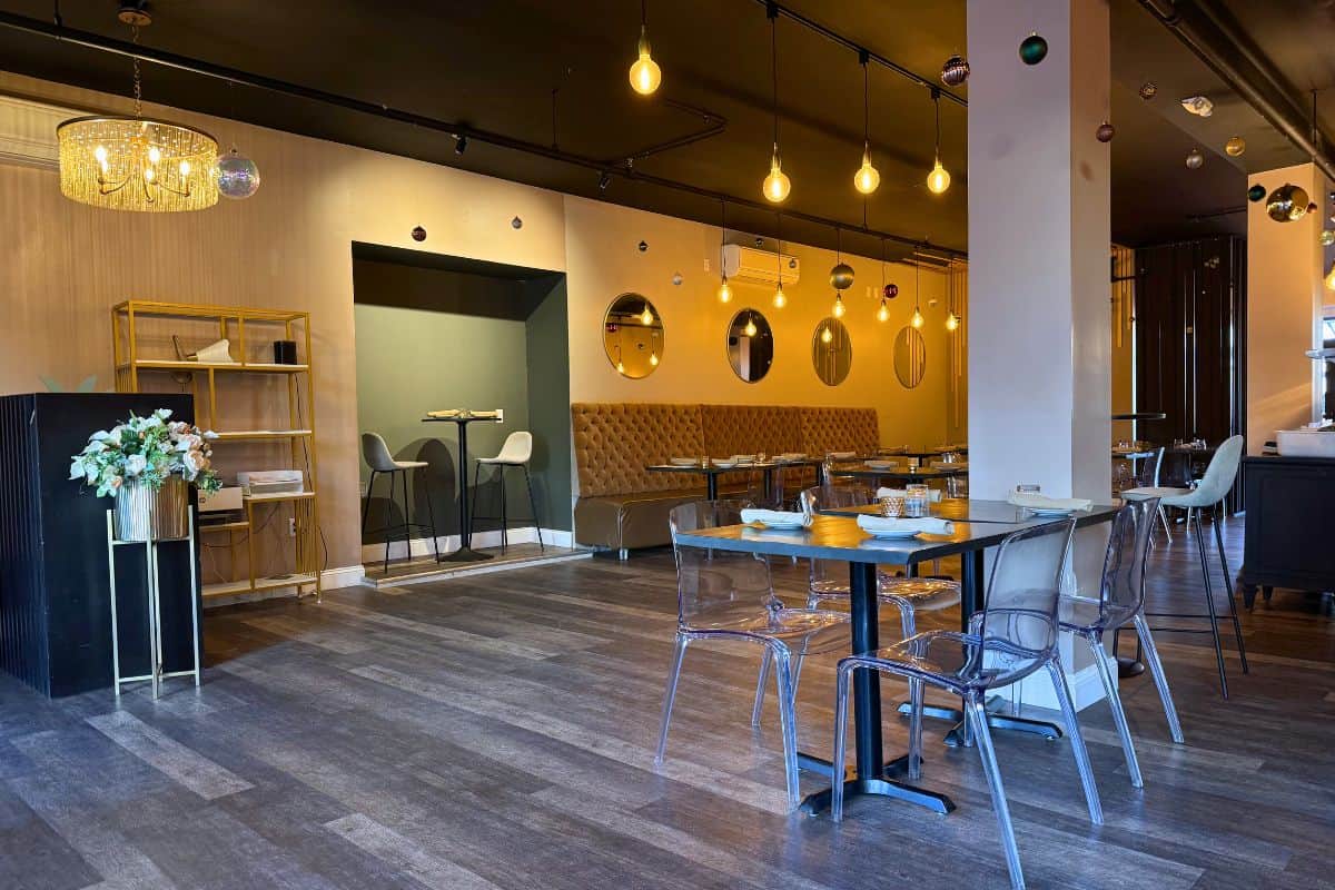 Modern restaurant interior with wooden floors, clear plastic chairs, tables set with dishes, wall mirrors, decorative lights, and a floral arrangement on a black stand.