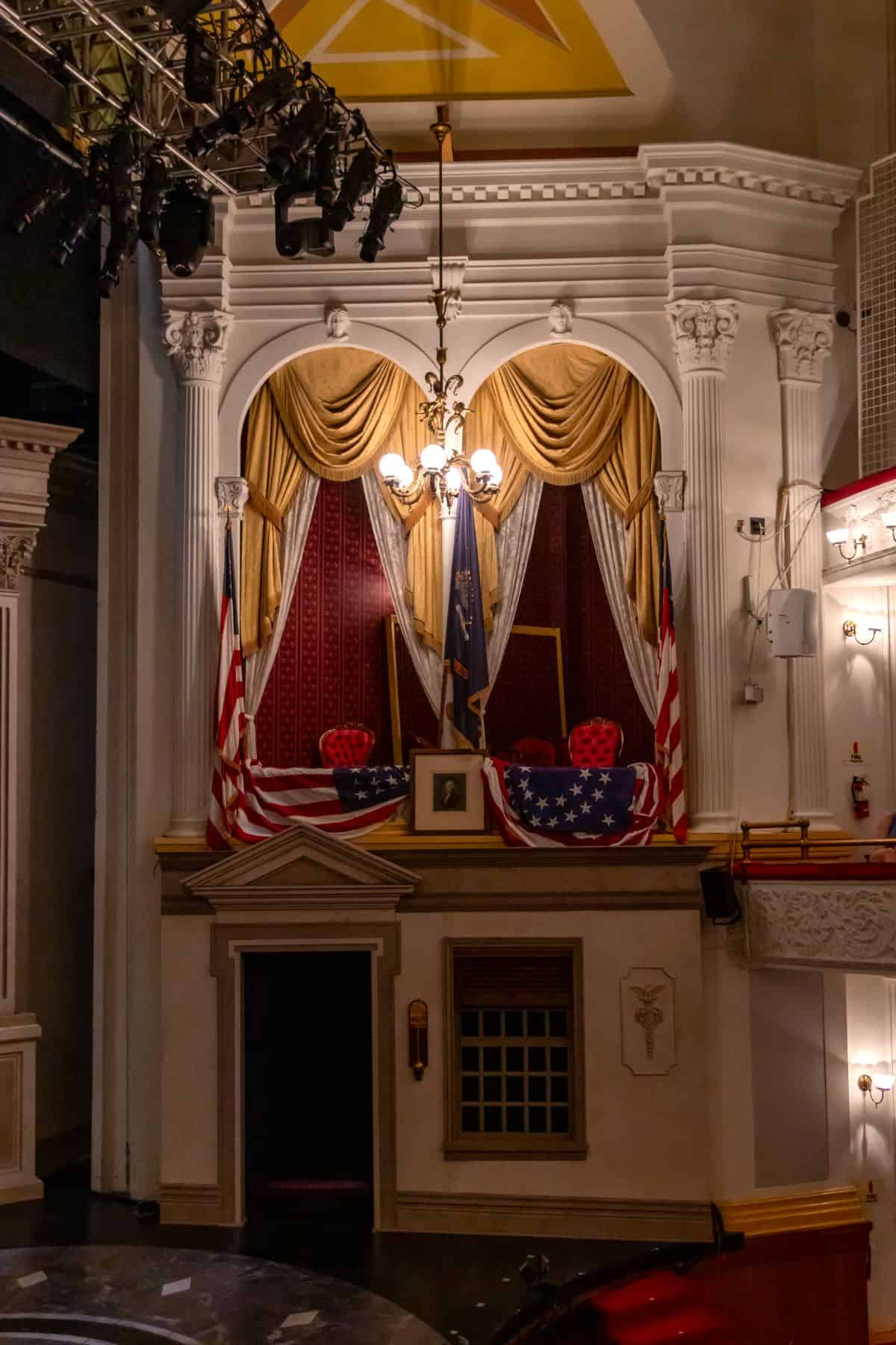 A theater box draped with American flags and gold curtains, featuring two chairs and a chandelier, in a historic setting.