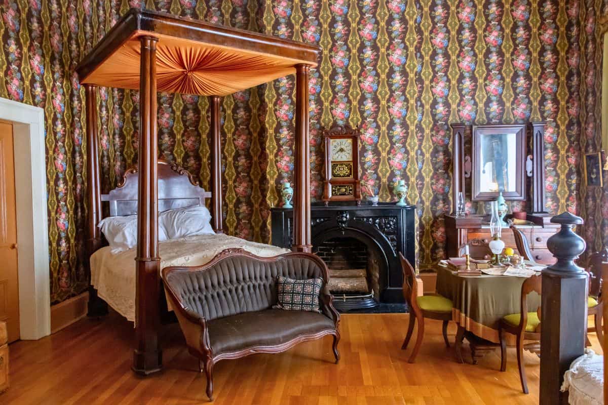 A vintage bedroom with a canopy bed, ornate wallpaper, a small sofa, a black fireplace, a dining table with chairs, and wooden floors.