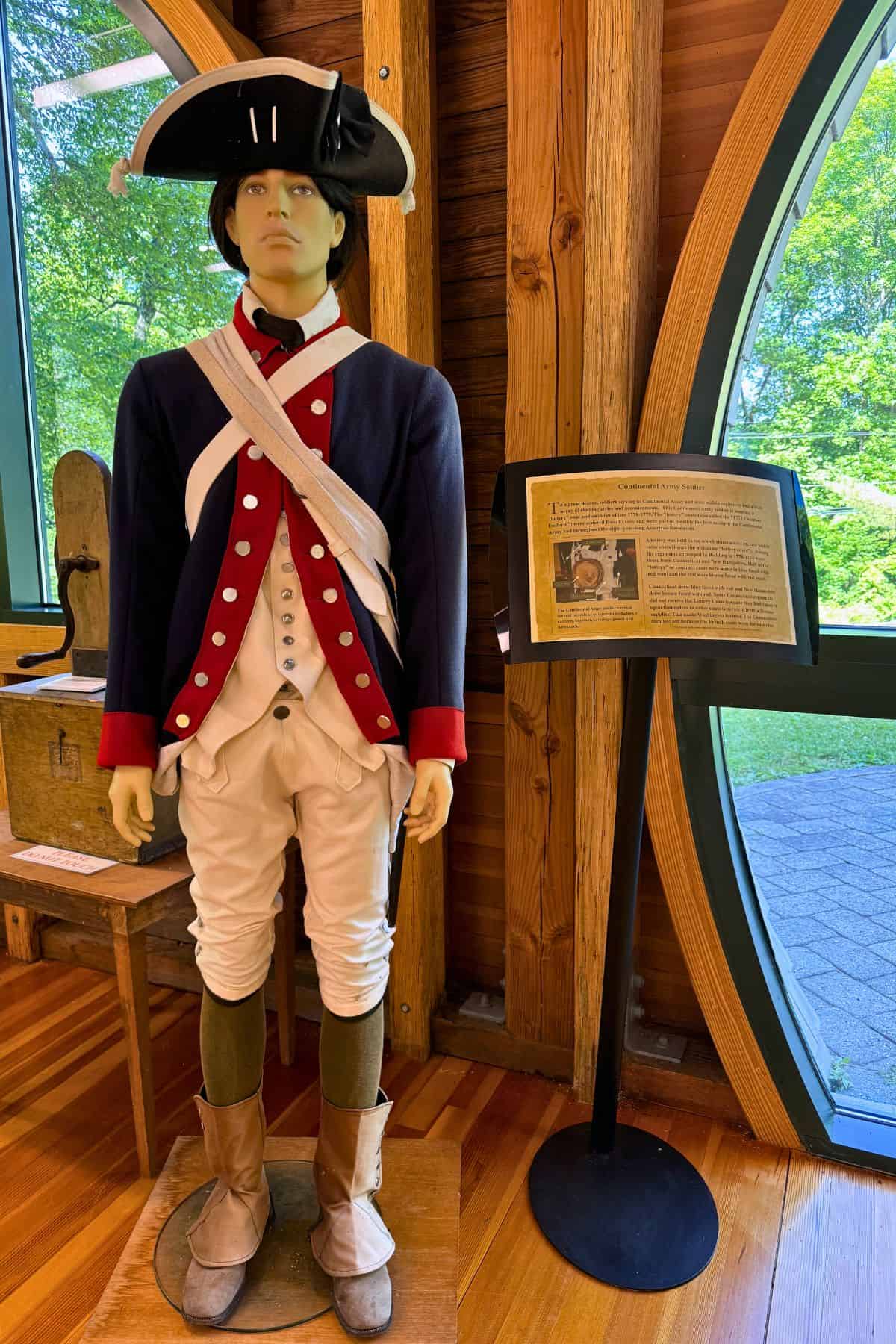 A mannequin dressed in a historical Revolutionary War soldier uniform stands indoors beside an informational display sign and a large window.